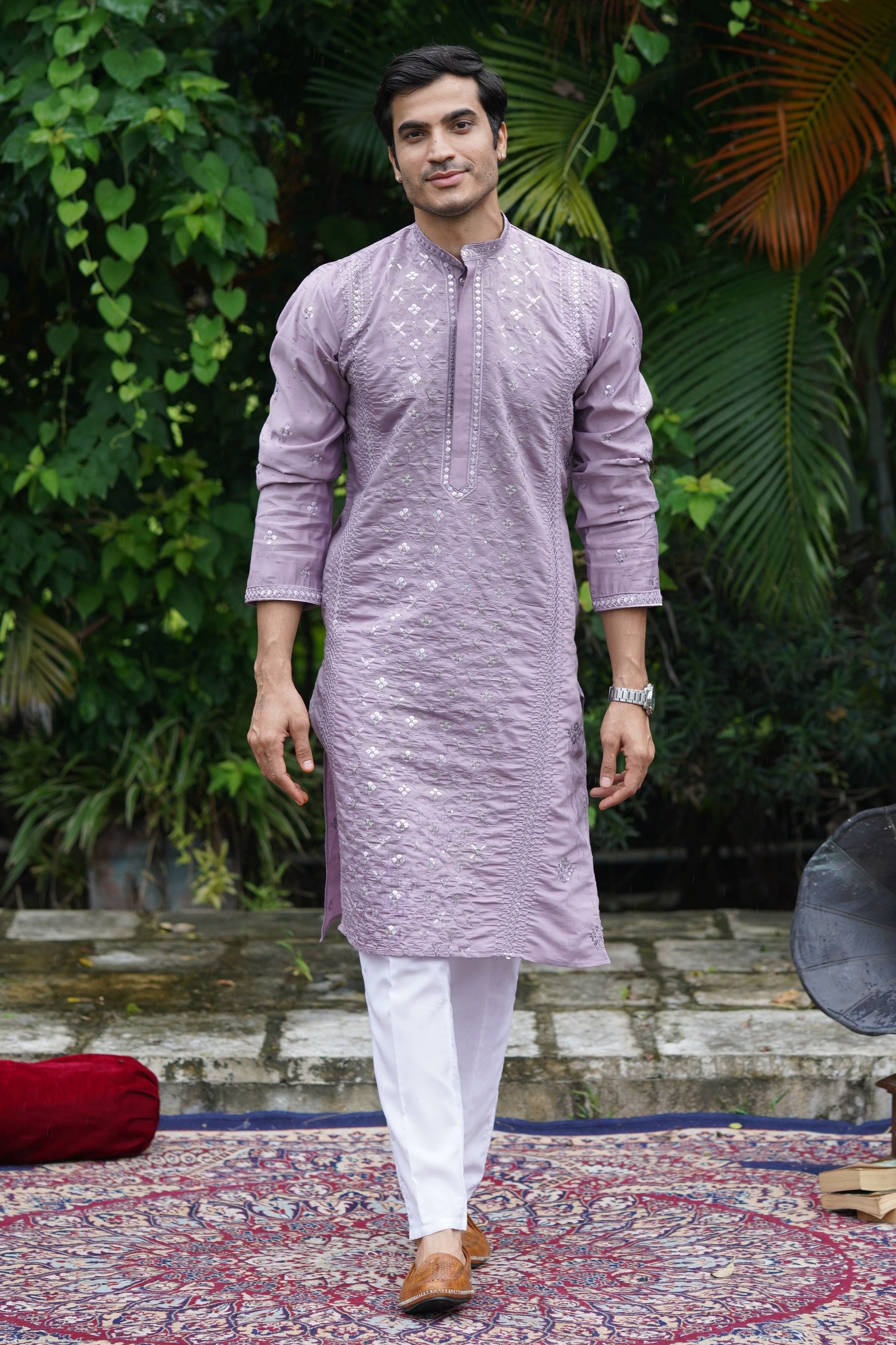 Person wearing a light purple traditional outfit standing in a garden with greenery and a decorative rug.